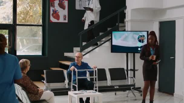 歩行フレームを持つ高齢者の待機エリアに座って 女性に専門医と医療相談に参加する前に話をする 病院の受付ロビーで待っている高齢者 — ストック動画