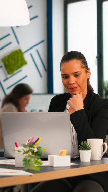 Dizüstü bilgisayarda yazı yazan mutlu İspanyol bayan, iş yerinde masa başında oturup kahve içerken arka planda çalışan çeşitli meslektaşlar. Yeni finans projesi planlayan çok ırklı iş arkadaşları.