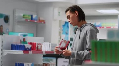 Eczanede ilaç kutusunu kontrol eden Asyalı genç adam ilaç ve takviye malzemesi almadan önce paket bilgilerini okuyor. Eczane raflarındaki vitamin ilaçlarına bakan yetişkin..