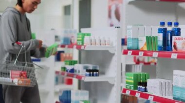 Genç adam eczane raflarından ilaç alıyor, reçeteli ilaç almak için sepetine ilaç koyuyor. Eczanede vitamin ve takviye kutularını kontrol eden Asyalı bir müşteri.