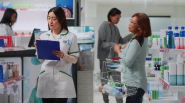 Asyalı uzman müşterilere kardiyoloji haplarıyla yardım ediyor, kadınlara ilaç veriyor ve tıbbî kaygıyı açıklıyor. Genç yetişkin hasta için eczane raflarından vitamin alıyor..