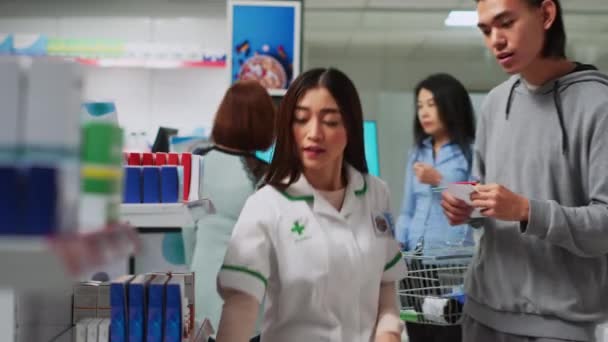 女性はサプリメントパッケージでアジア人男性を支援し 棚にビタミンの箱を説明します 薬局の小売店で処方薬について相談する男性の顧客 三脚ショット — ストック動画