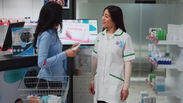 若い薬剤師はドラッグストアの女性を支援し アジアの顧客に薬や薬を与えるために処方紙を読んでいます 薬や薬の箱の話をしている人たちは — ストック動画