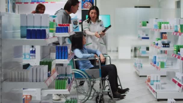 薬局の従業員は車椅子の女性に薬箱を与えます 障害のある人を助ける男性管理者 薬局の顧客やソーシャルワーカーに薬を与える薬剤師 — ストック動画