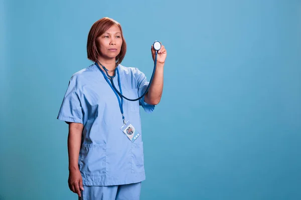 Elderly asian nurse in blue uniform holding medical stethoscope used for checkup consultation. Serious practitioner assistant working at healthcare facility, medicine service and concept