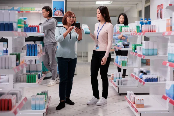 アジアの人々は薬局で薬や医療を見つけるためにスマートフォンを見て サプリメントやビタミンでお客様に薬の箱を与える医療専門家 — ストック写真