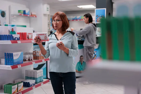 アジアのクライアントは ドラッグストアから処方薬を購入するために循環器系薬箱を検討し 薬やサプリメントなどの製品でいっぱいの棚 医療用品を見ている女性 — ストック写真