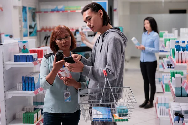 薬局で薬の治療を見つけるためにスマートフォンを見ている女性のアシスタントと若者 薬を購入する薬瓶をチェック 病気の男性の顧客を助けるドラッグストアの労働者 — ストック写真