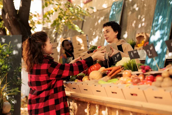 有機栽培の果物や野菜が入った段ボール箱を持つ若い女性の八百屋 地元の農家市場で健康的な自然野菜を販売するエプロン付きの笑顔のベンダー — ストック写真