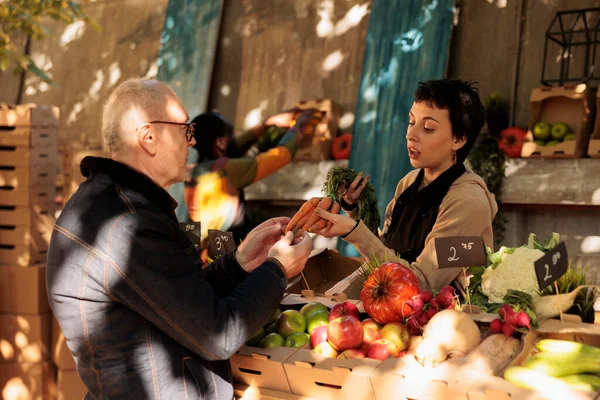 Female vendor showing fresh organic bunch carrots to mature man at farmers market. Small business owner standing behind fruit and vegetable stall selling healthy local home-grown produce to customer