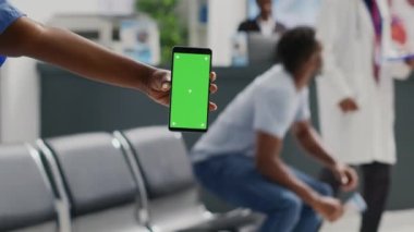 Hastane resepsiyonunda krom anahtar kullanarak elinde yeşil ekranlı cep telefonu tutan kadın hemşire. İzole edilmiş arka plan ve bekleme odasına bakan kişi.