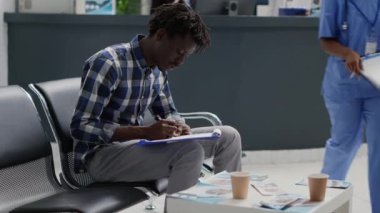 Afro-Amerikalı bir adam panoya rapor dolduruyor, doktorla randevuya başlamadan önce kayıt formu yazıyor. Bekleme alanında tıbbi dosyaları kullanan bir erkek..