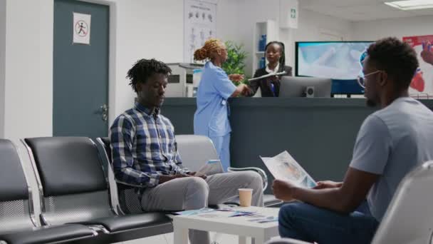 アフリカ系アメリカ人の男性は医療との協議に出席するのを待って 病院のレセプションに座っている 保健所で病気を治すための専門医との試験予約を持つ待機地域の若い大人 — ストック動画