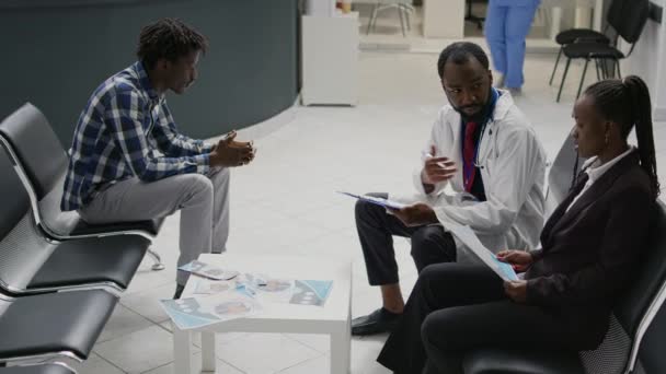 保健センターのロビーでアフリカ系アメリカ人女性の専門医に相談し 病気や医療について説明する 待機領域で医療アドバイスを与える専門家に耳を傾ける若い患者 — ストック動画