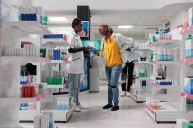 Eczane danışmanı alıcıya sırt ağrısı ilacı seçmesinde yardımcı oluyor. Sırtı ağrıyan Afrikalı Amerikalı kadın müşteri eczane koridorunda duruyor, alışveriş yapıyor, eczacıyla konuşuyor.