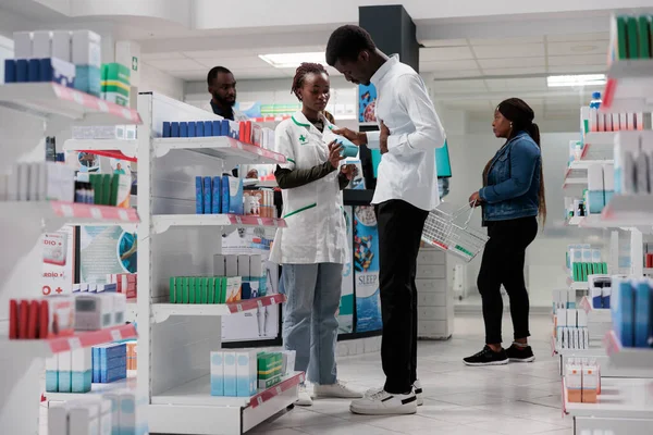 Customer buying stomachache medication, pharmacy consultant and buyer talking. African american man client with stomach pain standing in drugstore, shopping, chatting with pharmacist