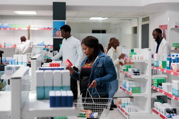 アフリカ系アメリカ人のバイヤーは薬局で心臓薬を選び 指示を読み 薬局の通路に立っています ビタミンを購入する女性の顧客 背景に薬剤師 すべての黒チーム — ストック写真
