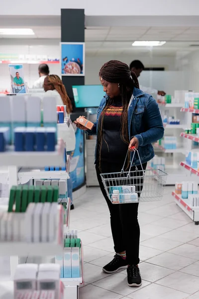 African american woman choosing sunscreen in pharmacy store, reading instruction on sunblock cream package. Buyer checking skin care lotion in drugstore, holding shopping basket