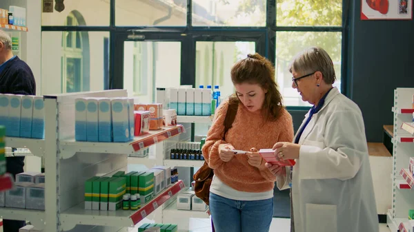 処方紙の錠剤治療について薬剤師に尋ねる若いクライアント 医療疾患治療を探しています 薬局の棚で薬やビタミンを探している女性 — ストック写真