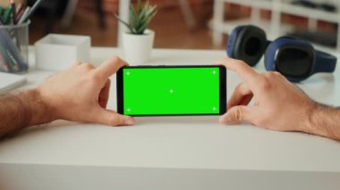 Genç yetişkin, akıllı telefondaki yatay yeşil ekran görüntüsünü analiz ediyor, masasında oturuyor, fotokopi alanı olan izole edilmiş model arka planına bakıyor. Krom anahtarlı telefonu tutmak. Kapatın. Üçayak atışı.
