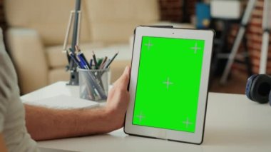 Ofis çalışanı dikey olarak yeşil ekranlı dijital cihazı tutuyor, kroma anahtar görüntüsüne ve boş model şablonuna bakıyor. Masadaki fotokopi alanı arkaplanı analiz ediliyor. Kapat..