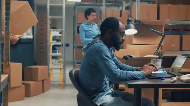 アフリカ系アメリカ人労働者は倉庫内の商品の品質を分析し 出荷のための商品注文を準備する 顧客に出荷するためにカートンボックスに消耗品や商品を入れて若い男 — ストック動画
