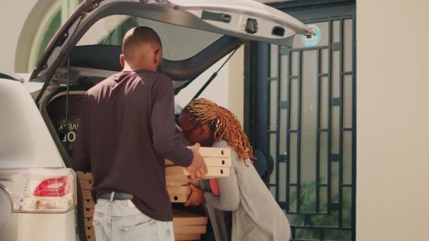 フロントドアの多様な顧客にピザパッケージの山を提供する女性のピッツェリア宅配業者 食品配達労働者は 車のトランクに食事パッケージのスタックで巨大な注文を与える 手持ち撮影 — ストック動画