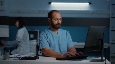 Hastane ofisinde gece geç saatlere kadar çalışırken kameraya gülümseyen pratisyen hemşire. Phyisician asistanı hasta raporunu analiz ediyor bilgisayarda tıbbi uzmanlık yazıyor.