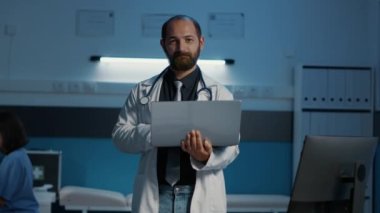 Tıbbi beyaz önlük ve steteskop giyen gülümseyen pratisyen doktor dizüstü bilgisayar tutuyor, sağlık hizmeti uzmanlığı yazıyor. Klinik personeli gece geç saatlere kadar hastane ofisinde çalışıyor. İlaç servisi.