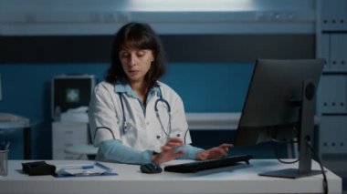 Specialist doctor standing at desk in hospital office working over hours at medical report while typing patient expertise on computer. Practitioner medic in white coat planning health care treatment