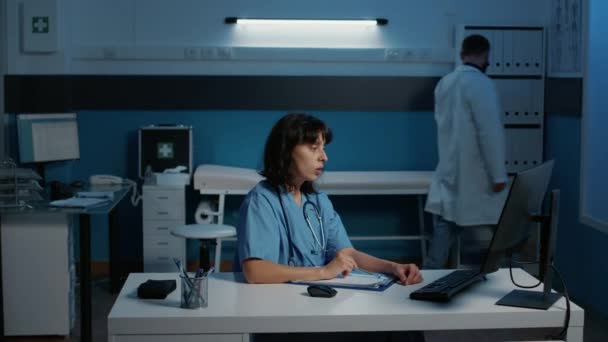 青い制服と聴診器を持つ医師の看護師は 病院のオフィスで夜遅くに医療治療で働く患者の病気の報告を分析します コンピュータに関する医療専門知識の確認アシスタント — ストック動画