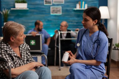 Hemşire huzurevindeki yaşlı kadın asistana yeni bir tedavi uyguluyor. Erkek hemşire, arka plandaki koltukta oturan yaşlı erkek hastayla ilgileniyor..