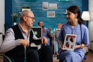 Uzman bir sağlık merkezinde hemşire, tekerlekli sandalyedeki yaşlı bir insana dijital tabletteki kalp görüntülerinin kalp fonksiyonlarını açıklıyor. Emekli adam huzurevinde tedavi görüyor..