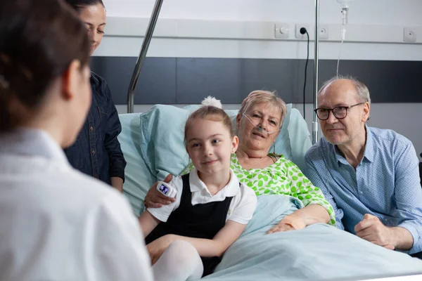 Elderly patient, family members listening attentively instructions of unrecognizable female health professional. Surgeon physician visiting grandmother bedridden in sanatorium room.