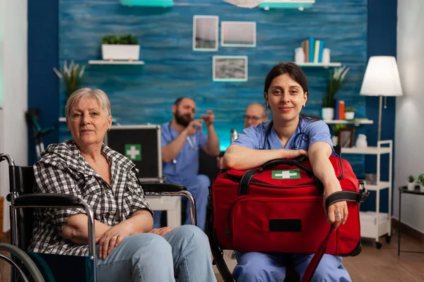Female healthcare assistant sitting in living room of nursing home next to elderly woman in wheelchair. Nurse holding bag of medical instruments to do physical checkup to patients in health center.