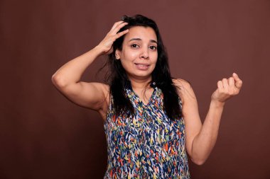 Düşünceli Hintli kadın, kafası karışık yüz ifadesi portresiyle ovalanıyor, garip pozlar veriyor, düşünceli hareketler yapıyor. Şaşkın gülümseyen, şüpheyle bakan, kameraya bakan, kahverengi arka planda orta ölçekli stüdyo görüntüsü olan