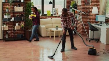 Afrika kökenli Amerikalı çift ev işleri ve ev işleri yapıyor, elektrikli süpürgeyle oturma odasını temizliyorlar. Mobilya ve raflardaki tozları paçavralar ve çamaşır çözeltileriyle süpüren bir kadın. El kamerasıyla..