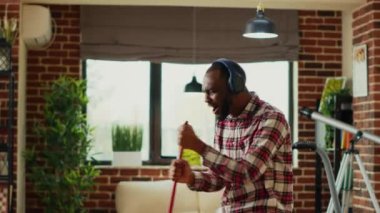 Genç ve neşeli bir yetişkin çamaşır makinesiyle yerleri paspaslıyor, dans ediyor ve kulaklıkla müzik dinliyor. Modern mutlu adam oturma odasını temizlerken, paspas kullanarak kiri temizlerken ve süpürürken eğleniyor..