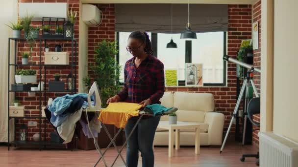 非洲裔美国女孩在冲浪板上用蒸汽做家务 休闲女友在客厅里熨烫洗衣衣服 年轻女人把衣服和衣服 家务活弄得整洁 — 图库视频影像
