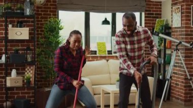 Mutlu adam ve kadın dans hareketleri yapıyor ve bahar temizliği yapıyor, yerleri paspas ve süpürgeyle temizliyor. Genç, gülümseyen bir çift ev işi yapıyor. El kamerasıyla..