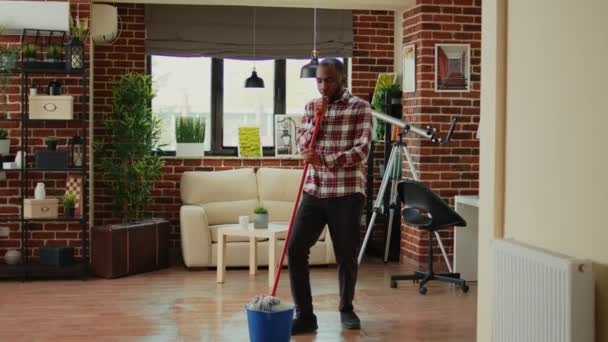 アフリカ系アメリカ人の男は家でモップとすべての目的クリーナーを使用して ほこりをきれいにするためにアパートの床をモッピング 汚れや混乱を掃除しながら 若い人が踊り 音楽を聴く — ストック動画