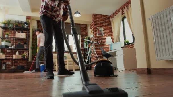 リビングルームでアフリカ系アメリカ人の男性が床を掃除し ガールフレンドと一緒にアパートを掃除します 掃除機を使用して若い大人と女性は すべての目的のクリーンな棚を拭く 三脚ショット — ストック動画