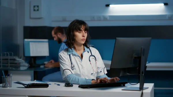 Portrait of physician doctor standing at desk in hospital office working over hours at health care treatment to help patient to cure disease. Woman medic wearing medical coat and stethoscope