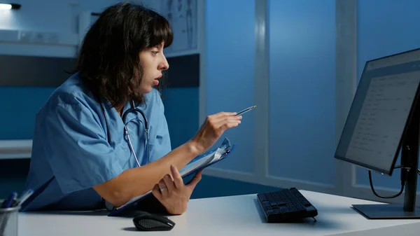 Practitioner assistant with blue uniform and stethoscope analyzing patient illness report working at health care treatment after hours in hospital office. Nurse checking medical expertise on computer
