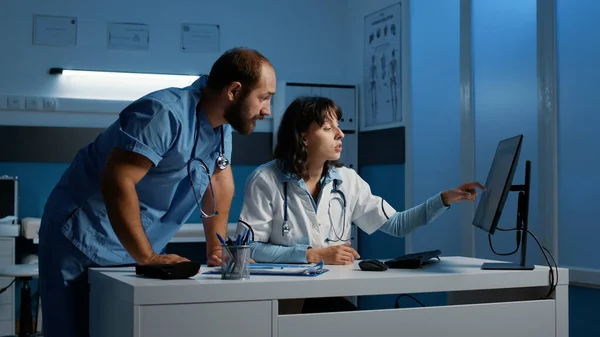 Clinical staff looking at computer monitor analyzing patient medical expertise while discussing medication treatment to help cure disease. Physician and nurse working night shift in hospital office