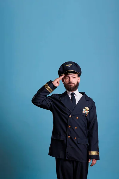 Serious aviation academy airplane aviator saluting, wearing uniform and hat front view portrait, plane pilot looking at camera. Middle age captain with airline wings badge on jacket