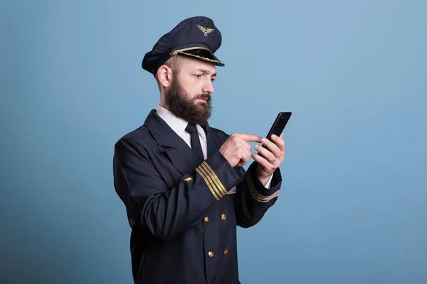 Plane aviator in aviation uniform watching online video on smartphone, holding mobile phone, looking at screen. Airlane captain standing, using telephone, studio medium shot