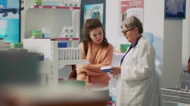Customer holding pills box and talking to pharmacist about treatment, looking at package of supplements. Young woman asking health specialist about vitamins and pharmaceutical products.