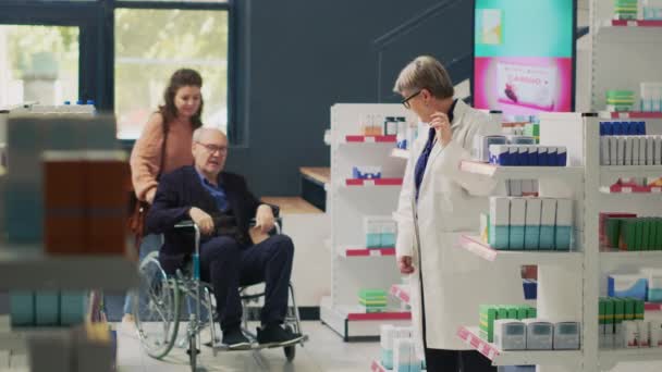薬局の薬局で薬剤師と話をしている高齢の顧客の車椅子のユーザーと管理人は 心臓病薬や薬について尋ねます 身体障害のある大人と介護者 三脚ショット — ストック動画