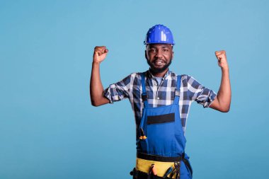 African american construction worker proud of developed arm muscles from the heavy work he performs daily. Builder wearing overalls and hard hat in studio shot against blue background.